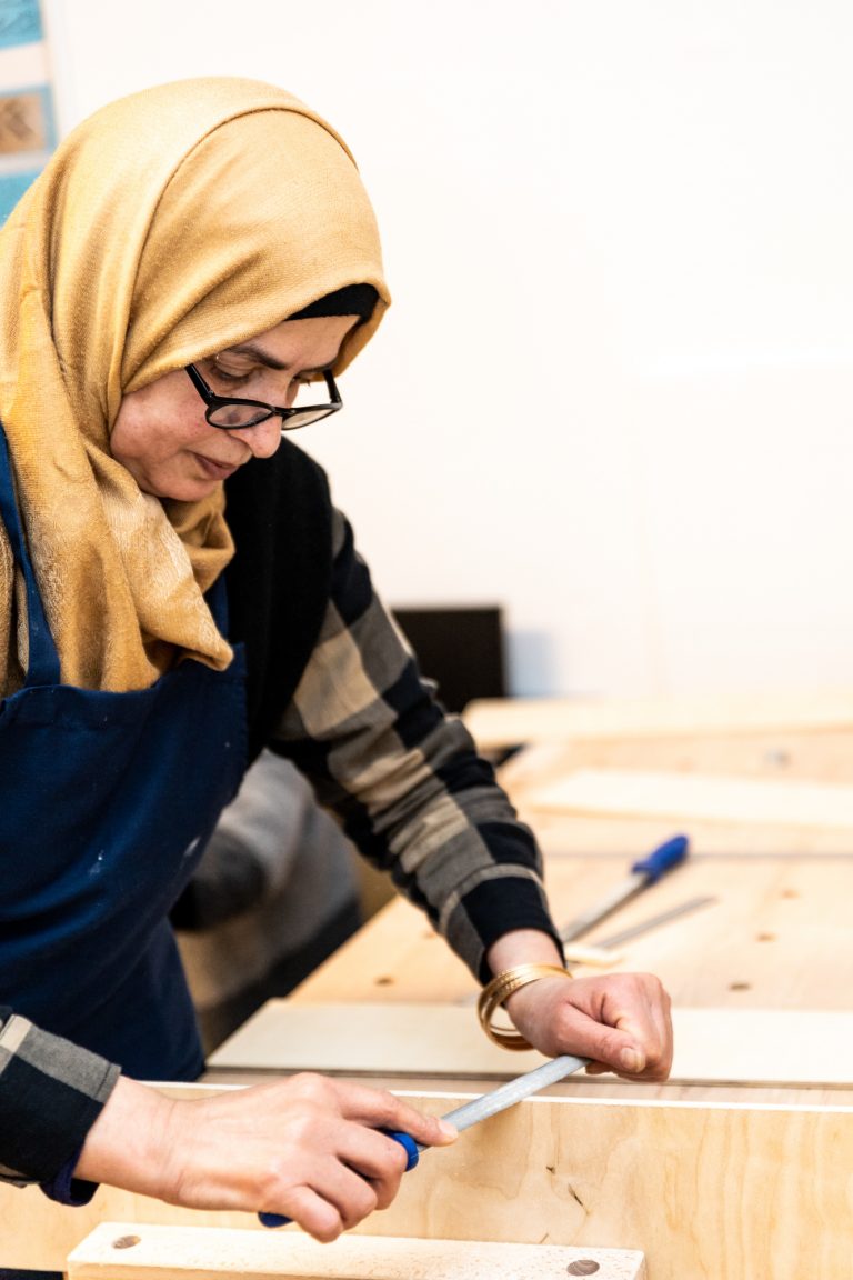 Eine Frau mit Kopftuch arbeitet mit einer Pfeile an einem Stück Holz auf einer Werkbank. Imagefoto für den Kommunalen Eigenbetrieb Leipzig / Engelsdorf (KEE).