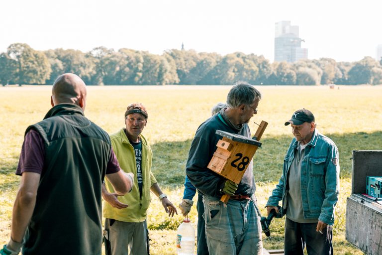 Vier Männer in Arbeitskleidung stehen an einer Wiese im Rosental in Leipzig. Eine hält einen Nistkasten für Vögel in der Hand, der an einem Baum angebracht werden soll. Imagefoto für den Kommunalen Eigenbetrieb Leipzig / Engelsdorf.