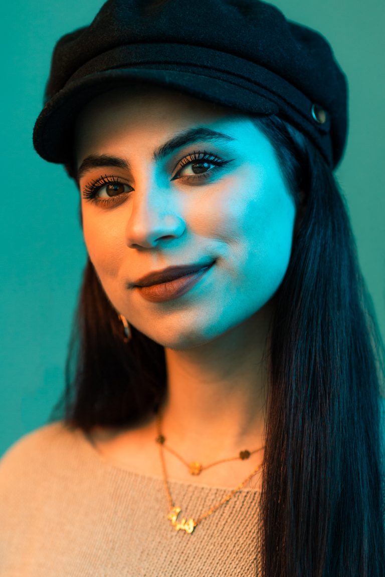 Portrait in Orange und Türkis. Portraitfoto einer jungen Frau mit Schirmmütze und dunklen Haaren, Ohrringen und einer Halskette.
