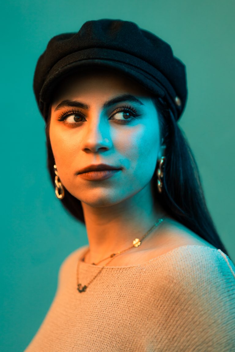 Portrait in Orange und Türkis. Portraitfoto einer jungen Frau mit Schirmmütze und dunklen Haaren, Ohrringen und einer Halskette.