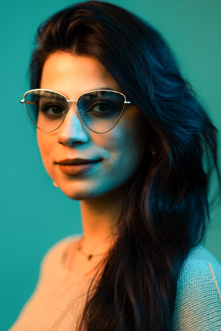 Portrait in Orange und Türkis. Eine junge Frau mit Sonnenbrille und dunklen Haaren schaut in die Kamera.