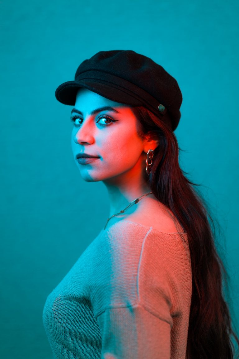 Portrait in rotem und blauem Licht. Portraitfoto einer jungen Frau mit Schirmmütze und dunklen Haaren, Ohrringen und einer Halskette.