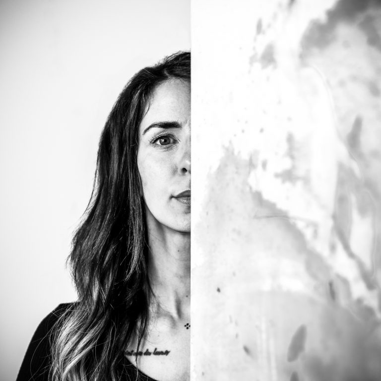 Portrait in Schwarz-Weiß. Es ist die Hälfte des Gesichts einer Künstlerin zu sehen. Sie schaut entschlossen in die Kamera. Die andere Gesichtshälfte wird von einem Gemälde verdeckt.