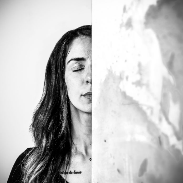 Portrait in Schwarz-Weiß. Es ist die Hälfte des Gesichts einer Künstlerin zu sehen. Ihr Auge ist geschlossen. Die andere Gesichtshälfte wird von einem Gemälde verdeckt.