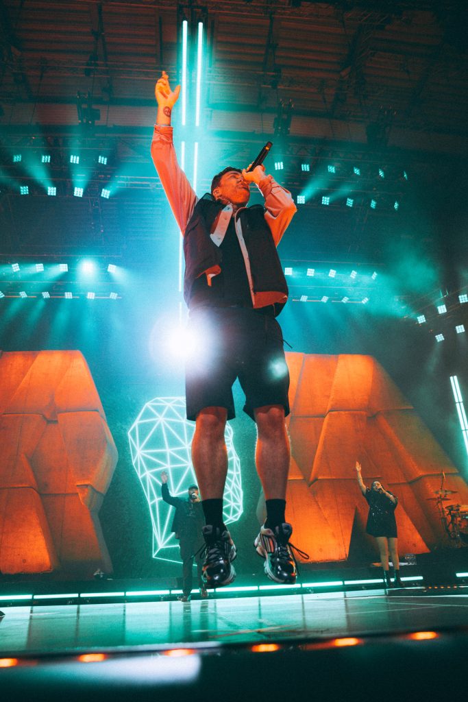 Concert photography Leipzig: Marteria im Sprung mit erhobenem Arm, türkises Bühnenlicht, Backgroundsänger im Hintergrund