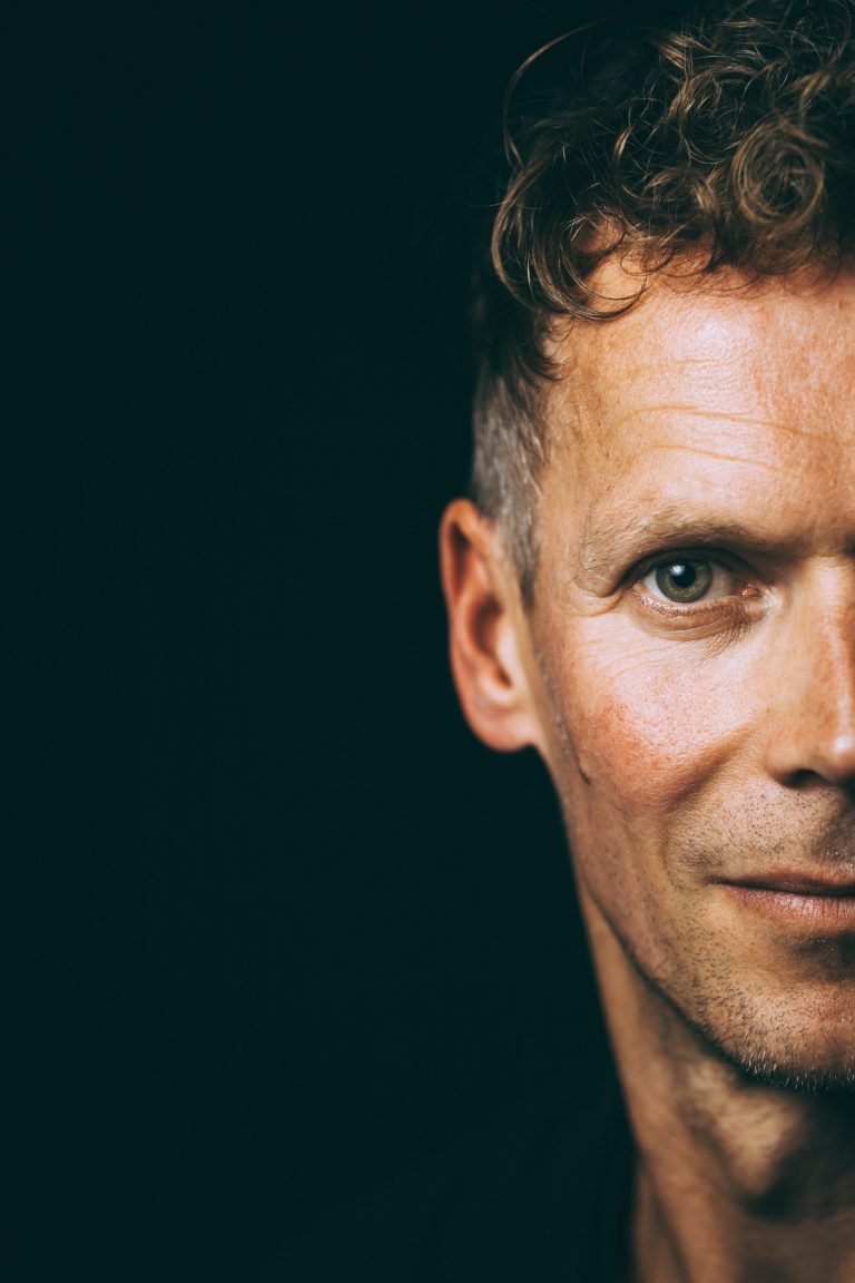 Selbstbewusster Headshot eines Mannes im dunklen Studio, nur das halbe Gesicht ist sichtbar, entschlossener Blick, Low-Key-Portrait